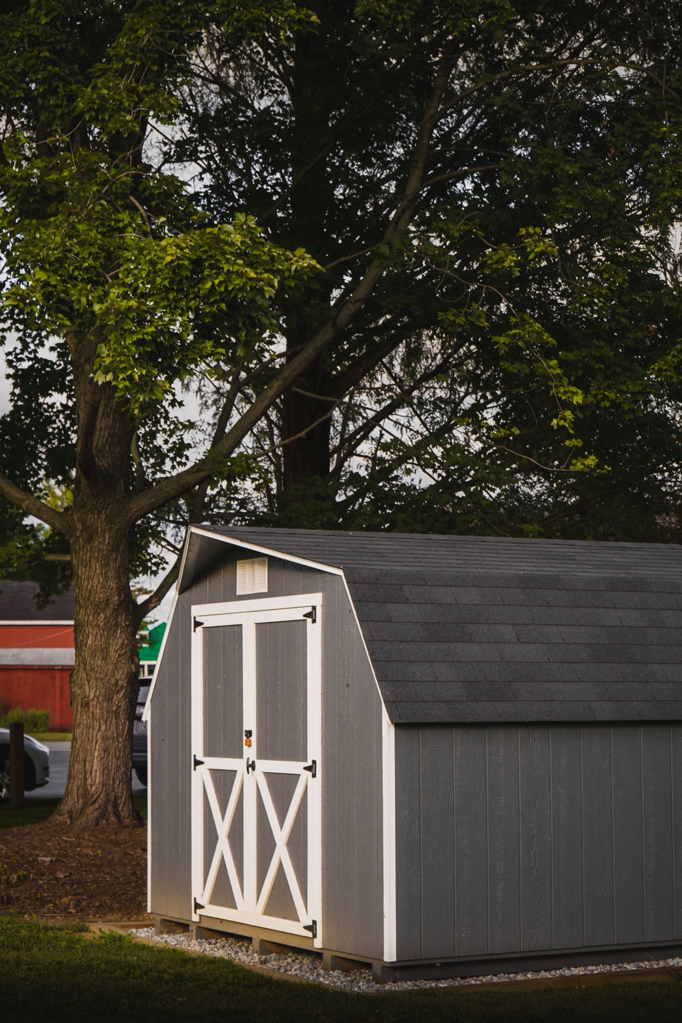 Gray gambrel-roofed backyard storage shed with white trim under a large shade tree, cooling a shed diy project